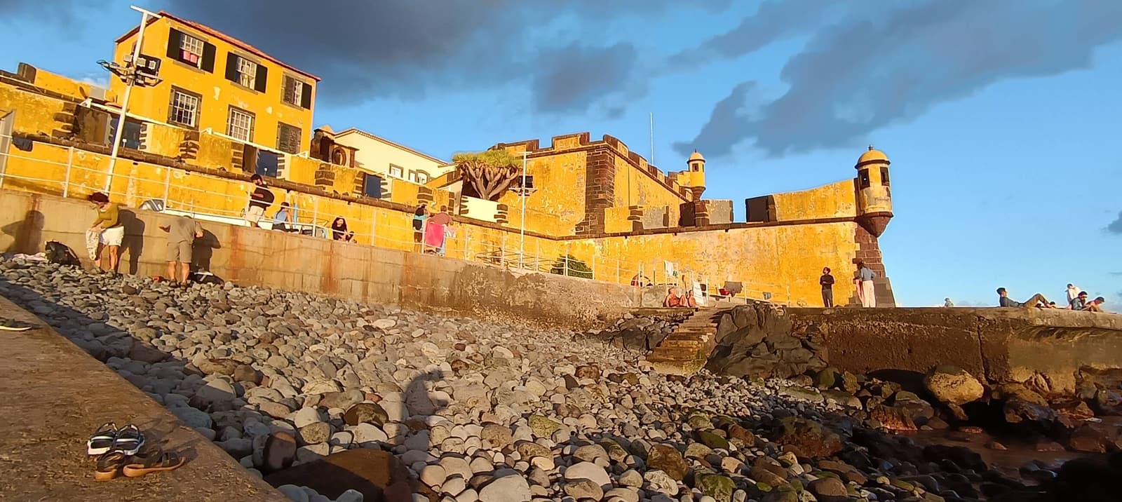 The yellow walls of Forte de São Tiago above a pebble beach in Funchal at golden hour, people sitting on the stones