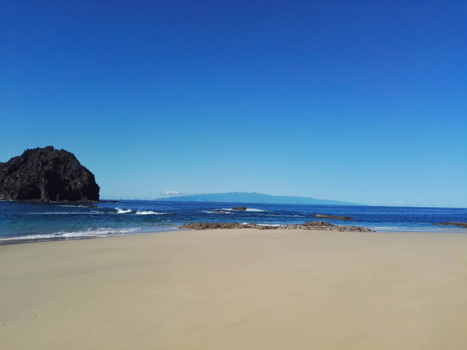 A sandy beach with a dark volcanic rock outcrop and distant island silhouette on the horizon, at Prainha, Madeira